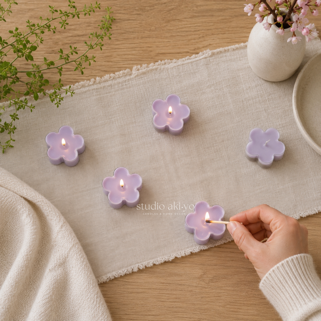 Flower-shaped candles being lit on a table with a hand, plants, and a vase in the background.