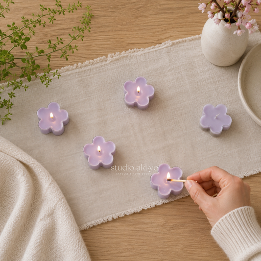 Flower-shaped candles being lit on a table with a hand, plants, and a vase in the background.