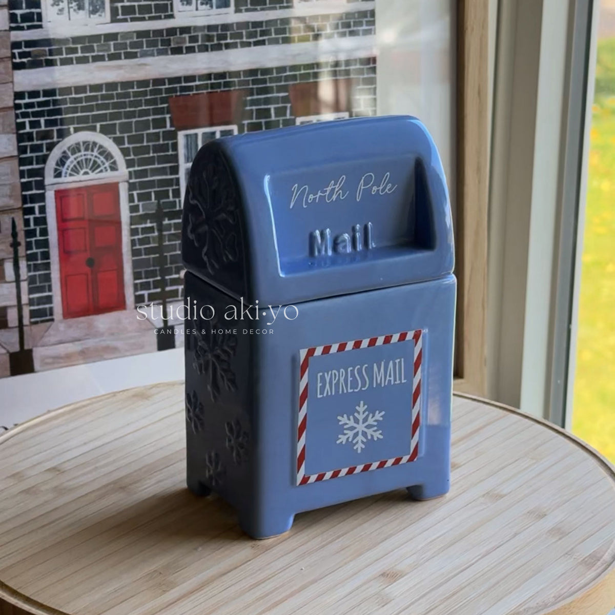 A blue ceramic North Pole Express Mail box with embossed details, sitting on a light wooden table in front of a window with a miniature house backdrop.