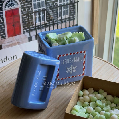A light blue square ceramic container labeled 'EXPRESS MAIL' with a matching lid placed beside it on a wooden table. The container is filled with pale green wax melts, and an open cardboard box of more wax melts sits in the foreground.