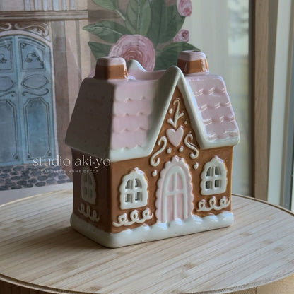 A gingerbread house-shaped ceramic container or candle holder with pink icing-style detailing, displayed on a round wooden surface in front of a softly blurred floral and architectural background.