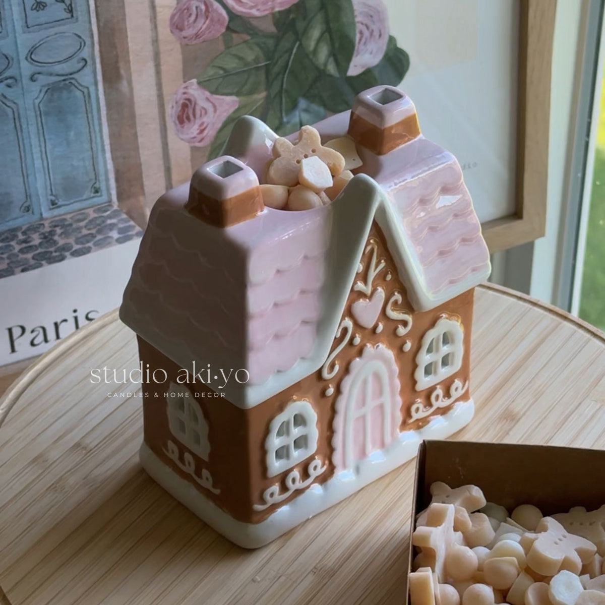 A gingerbread house-shaped candle holder or decorative candle tin with a pink roof and white trim, placed on a wooden surface. A small box of cream-colored wax pieces sits beside it, and a framed floral print is visible in the background.