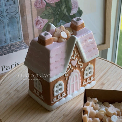 A gingerbread house-shaped candle holder or decorative candle tin with a pink roof and white trim, placed on a wooden surface. A small box of cream-colored wax pieces sits beside it, and a framed floral print is visible in the background.