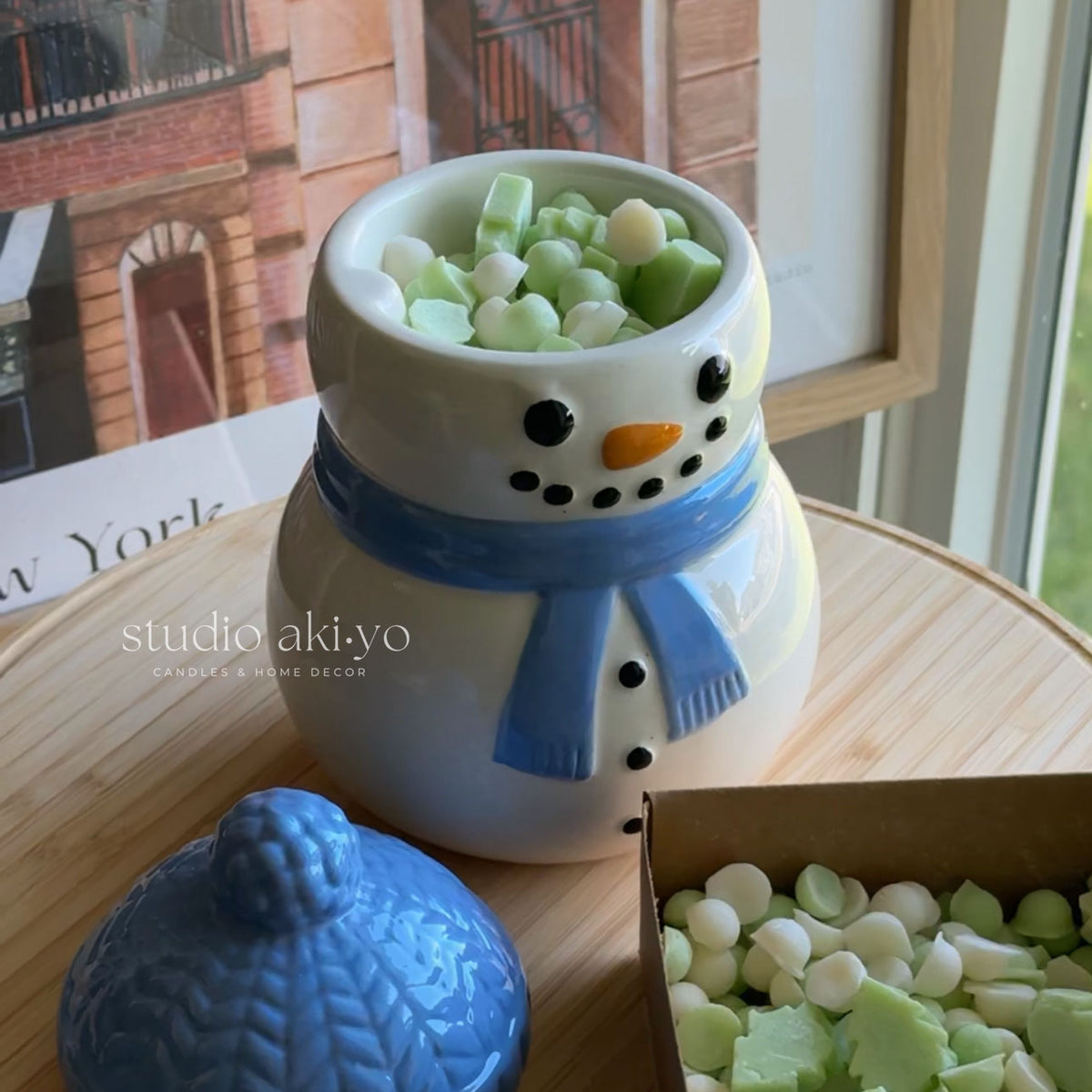 A ceramic snowman candle holder with a blue scarf and face details, filled with small green and white wax candles, shown on a wooden table with its blue lid beside it and a box of more candles nearby.
