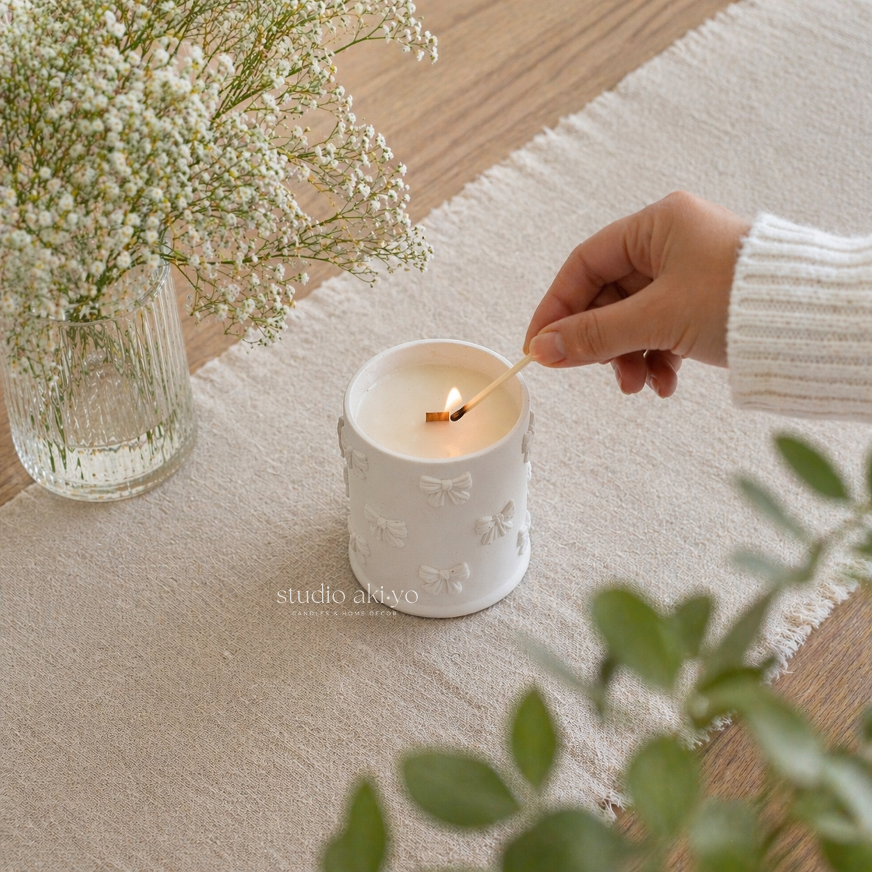 Hand lighting a white candle with floral design on a textured surface, next to a vase of flowers.