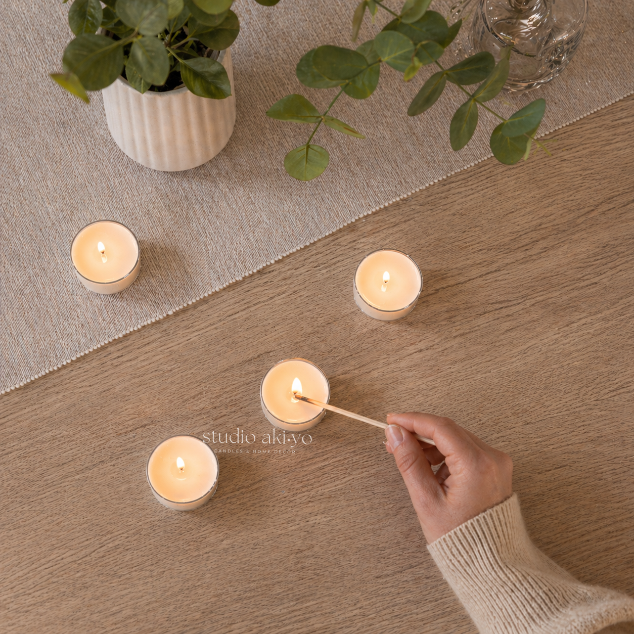 Hand lighting Studio Akiyo tealight candles on a wooden surface with a potted plant in the background