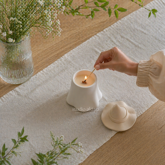 Person lighting a candle on a table with a vase of flowers and a hat in the background.