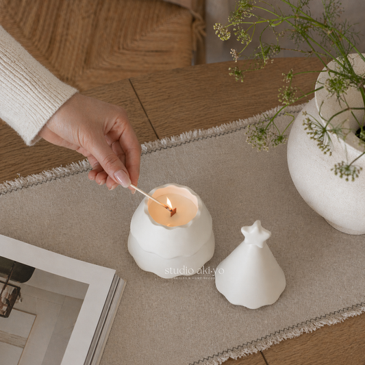 Person lighting a candle in a white holder on a wooden table with a vase of flowers and a book in the background.