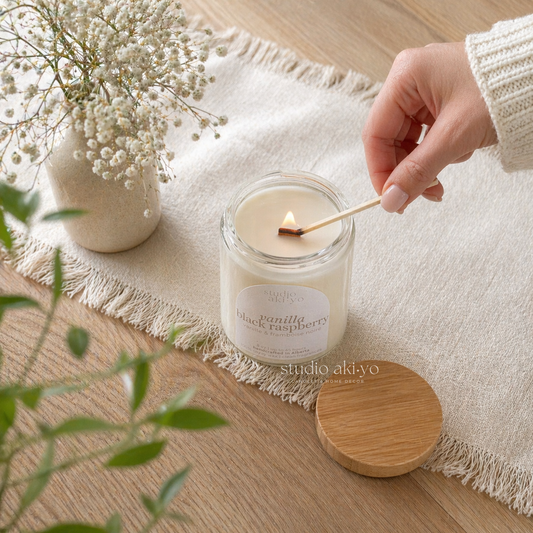 Hand lighting a Studio Akiyo candle with a match on a wooden surface with a vase of flowers and greenery.