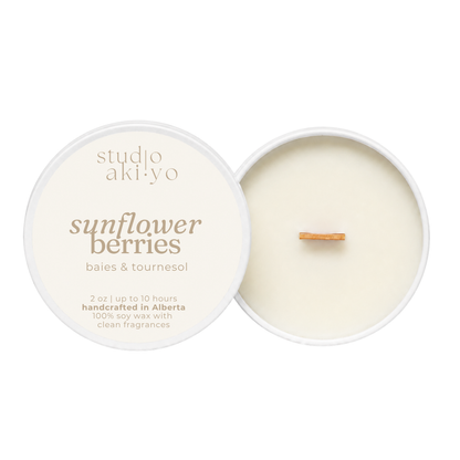 A round soy wax candle shown open with the cream-colored lid beside it on a white background. The lid reads 'studio aki·yo' and 'sunflower berries' with smaller text 'baies & tournesol' and '2 oz | up to 10 hours handcrafted in Alberta 100% soy wax with clean fragrances.