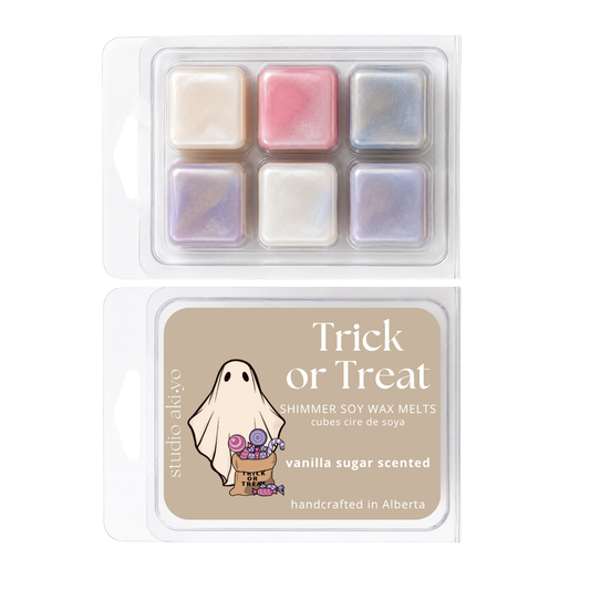 Two clear plastic clamshell packages of Studio Akiyo 'Trick or Treat' shimmer soy wax melts. The top package shows six square wax cubes in pastel shades of cream, pink, and iridescent blue.