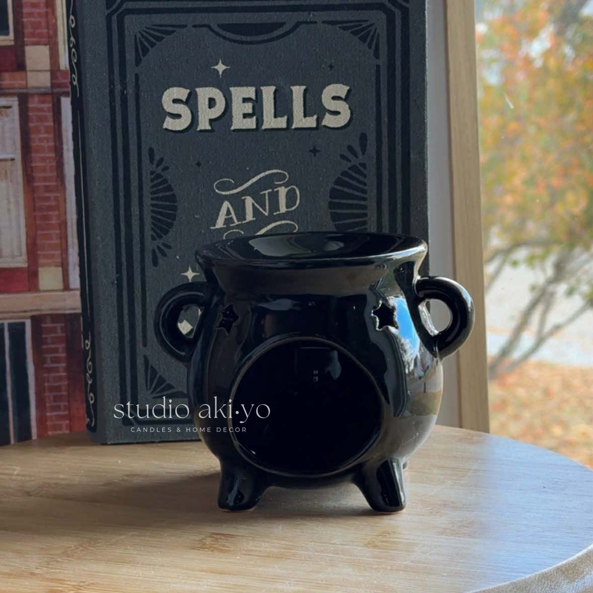 Black ceramic wtichy cauldron wax warmer with a lit candle inside, surrounded by decorative items on a table.
