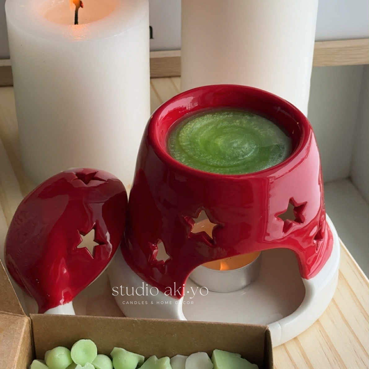Red ceramic wax warmer with green wax melts next to a lit candle on a wooden surface.