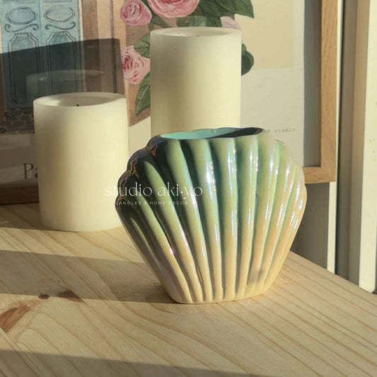 Decorative ceramic vase with ribbed design on a wooden surface, with candles and a floral painting in the background.