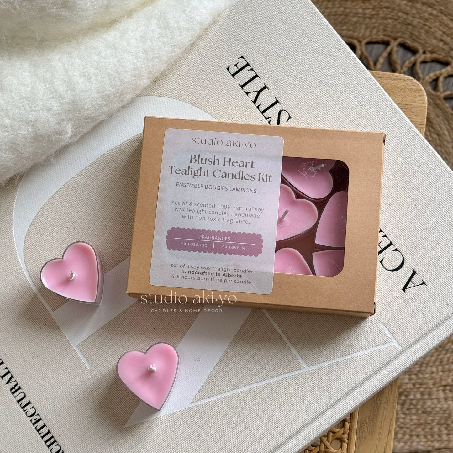 A set of pink heart-shaped tealight candles presented in a box, with four candles visible outside the box, placed on a wooden surface with a book and a dried plant in the background.