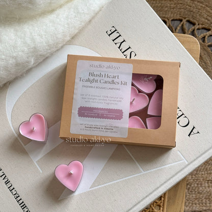 A set of pink heart-shaped tealight candles presented in a box, with four candles visible outside the box, placed on a wooden surface with a book and a dried plant in the background.