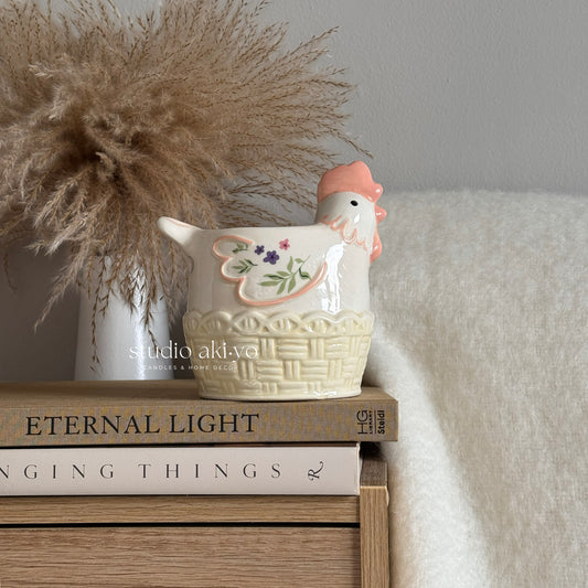 Ceramic chicken-shaped wax melt burner on a stack of books with decorative elements.