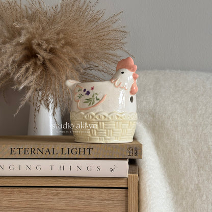 Ceramic floral hen chicken-shaped wax melt burner on a stack of books with decorative elements.
