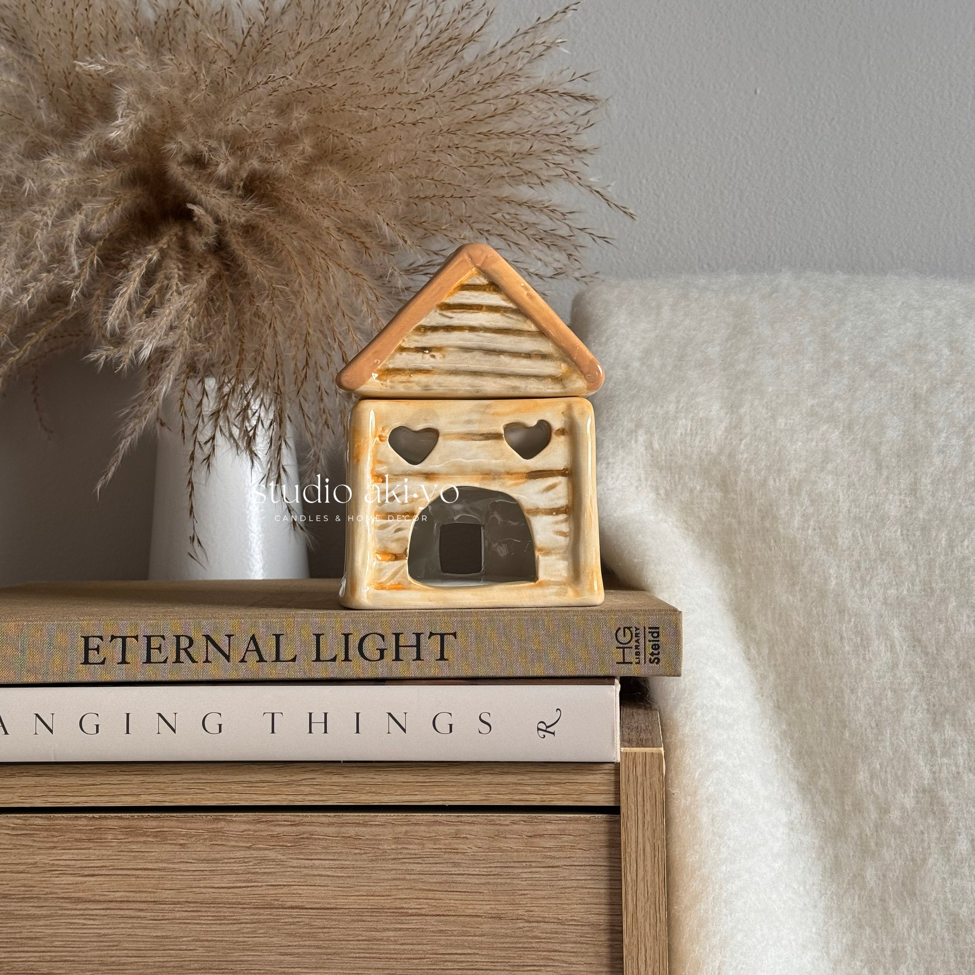 Decorative house-shaped lantern on books with a vase of dried pampas grass in the background.