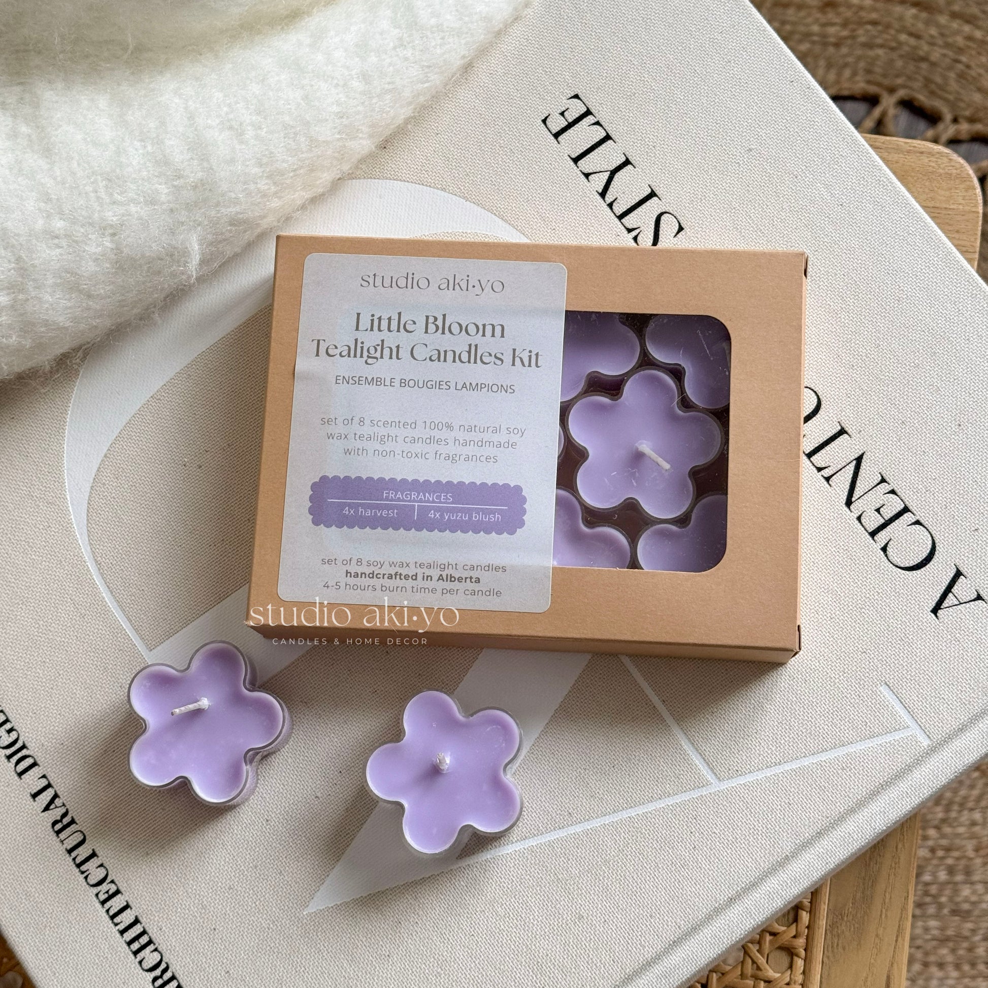 A set of purple flower-shaped tealight candles from Studio Akiyo's Little Bloom collection, displayed on top of a book with the product packaging in the background.