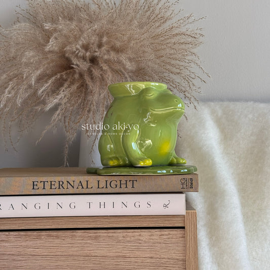 Green ceramic frog sculpture on books with decorative pampas grass and white fabric in the background