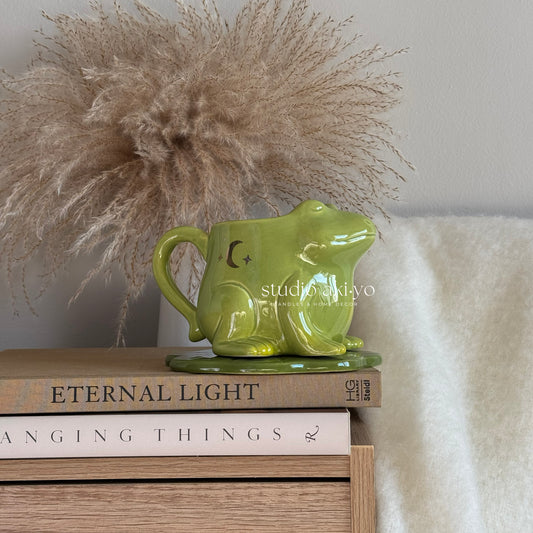 Green ceramic frog-shaped object on books with decorative pampas grass and soft fabric in the background