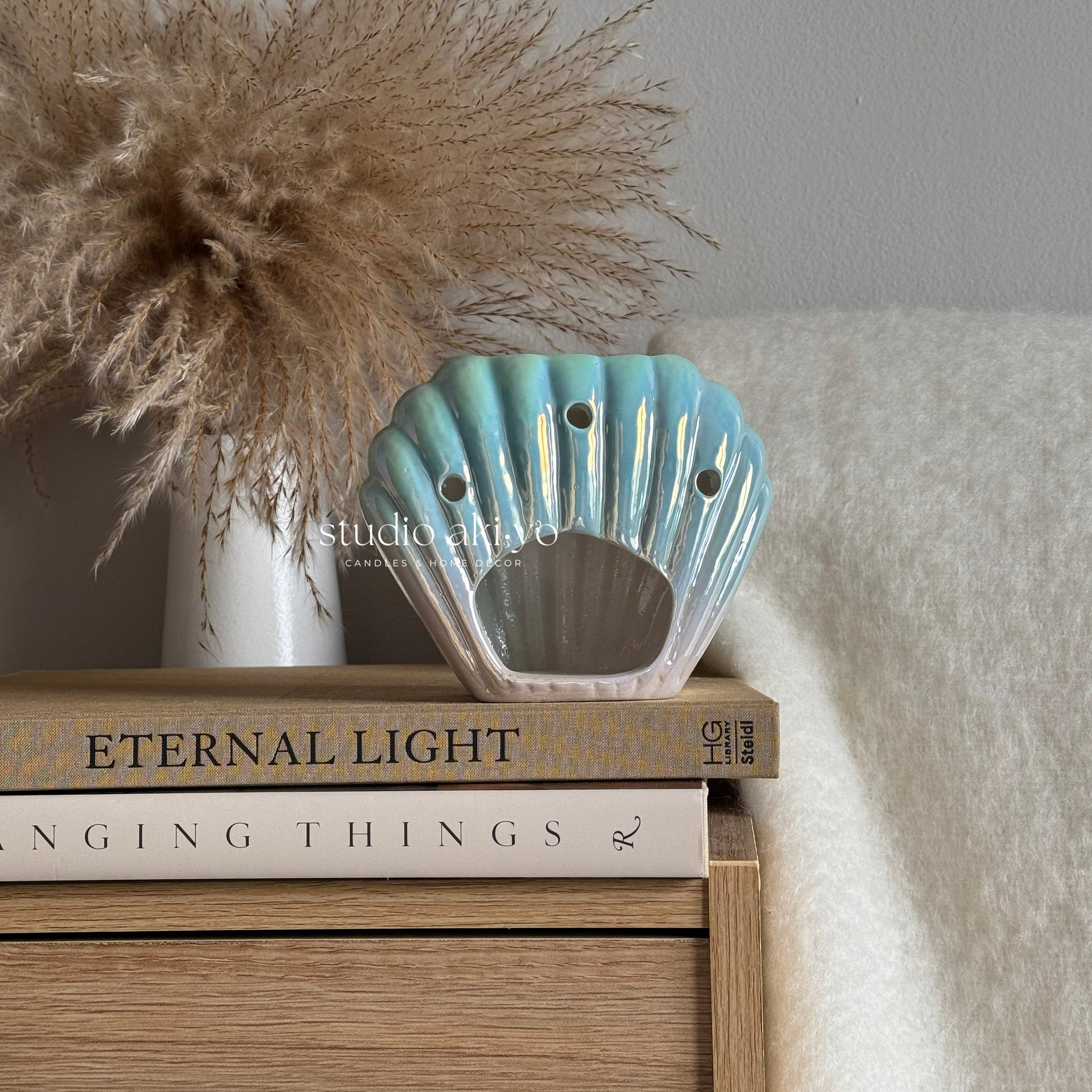Decorative shell-shaped wax melt burner on books with a vase of dried pampas grass in the background.