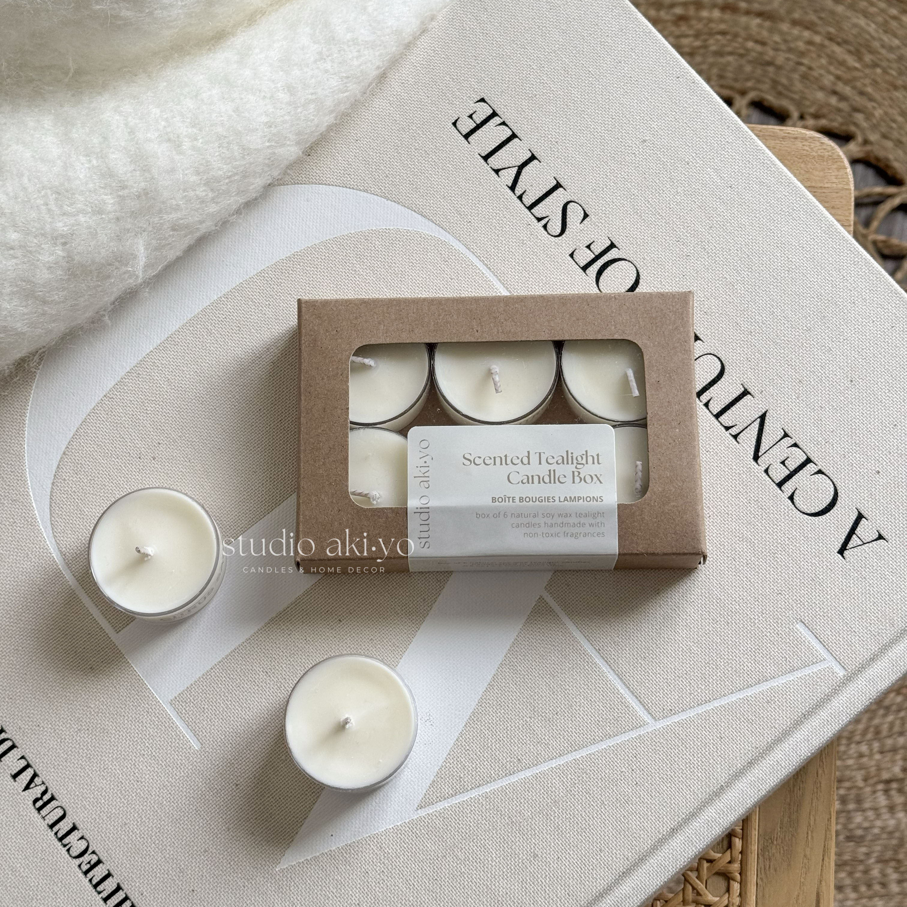 A box of six white tealight candles with a 'Studio Akiyo' label on it, displayed next to a lit candle on a white plate, with a white vase and decorative items in the background.