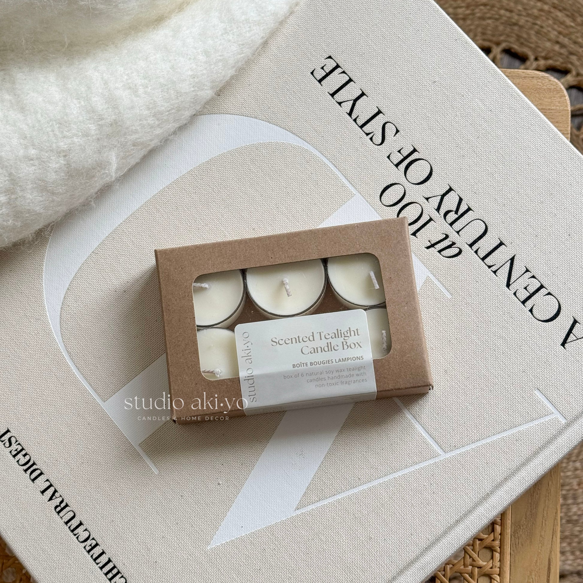 A box of six white tealight candles with a 'Studio Akiyo' label on it, displayed next to a lit candle on a white plate, with a white vase and decorative items in the background.