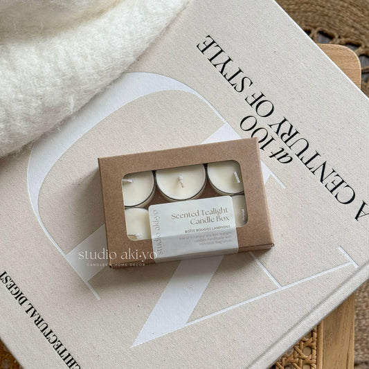 A box of six white tealight candles with a 'Studio Akiyo' label on it, displayed next to a lit candle on a white plate, with a white vase and decorative items in the background.