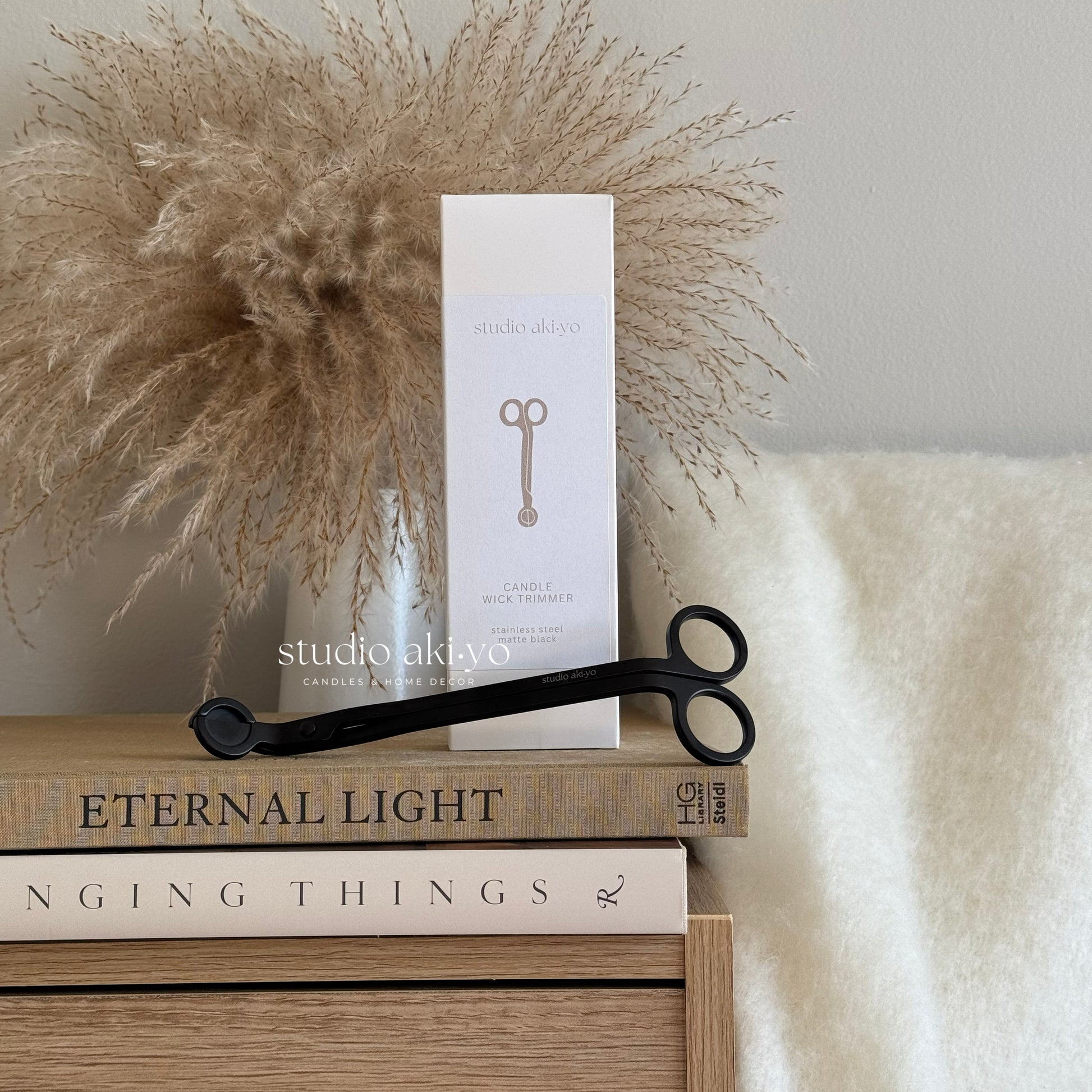 Candle box with scissors on books next to decorative pampas grass against a neutral background