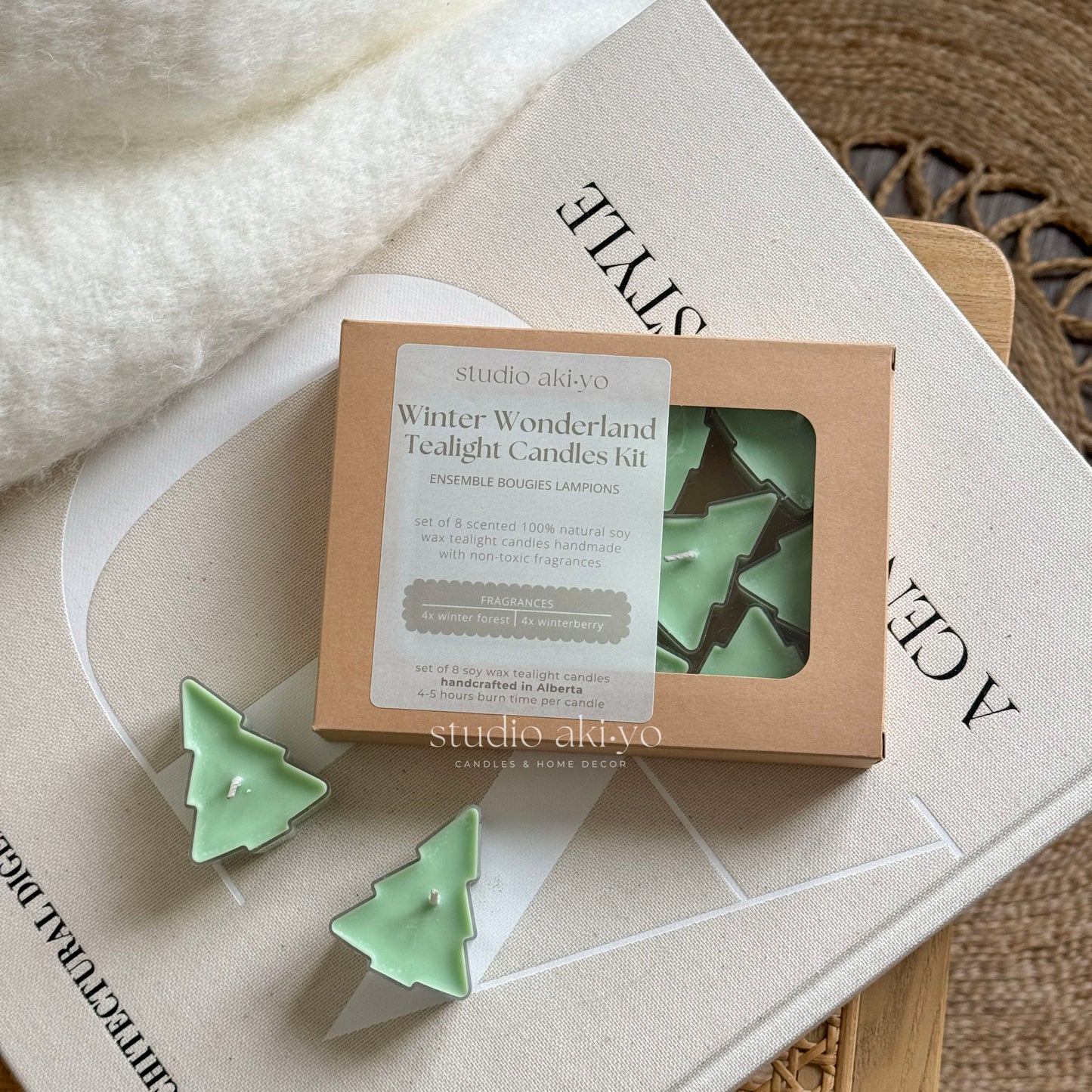 A set of green tree-shaped tealight candles presented in a box, with the product name 'Winter Wonderland Tealight Candles Kit' visible. The candles are displayed in a clearing surrounded by foliage and set on a wooden surface with books and decorative items in the background.