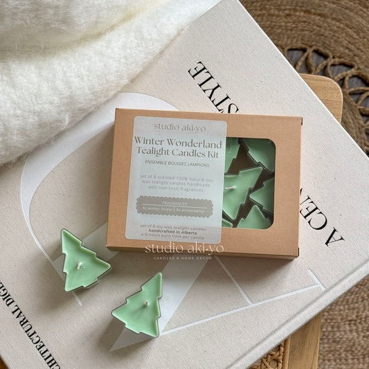 A set of green tree-shaped tealight candles presented in a box, with the product name 'Winter Wonderland Tealight Candles Kit' visible. The candles are displayed in a clearing surrounded by foliage and set on a wooden surface with books and decorative items in the background.