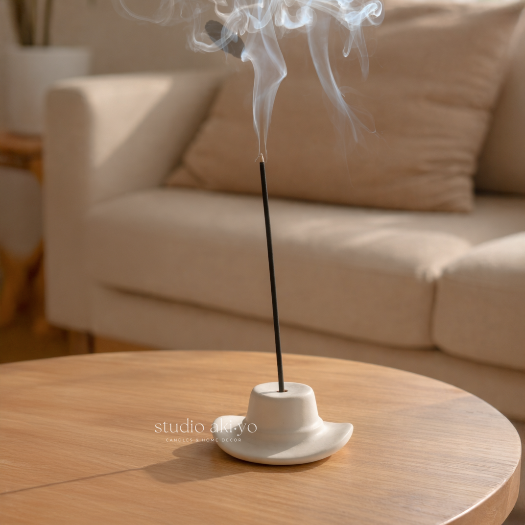 Incense stick with smoke on a white holder on a wooden table, blurred beige sofa in the background.